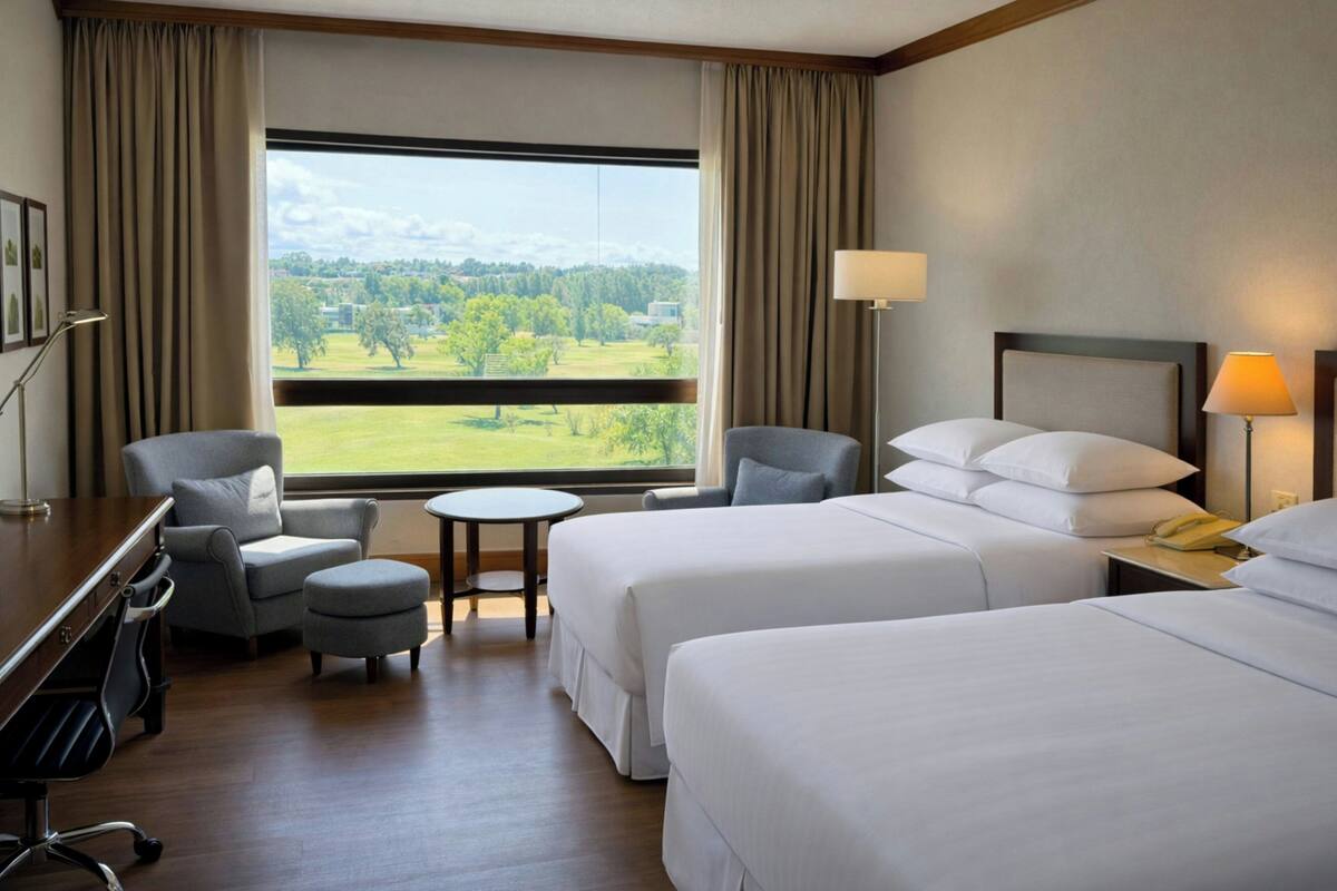 Habitación de hotel con dos camas, un escritorio, una silla, una mesita y vista a un paisaje verde.