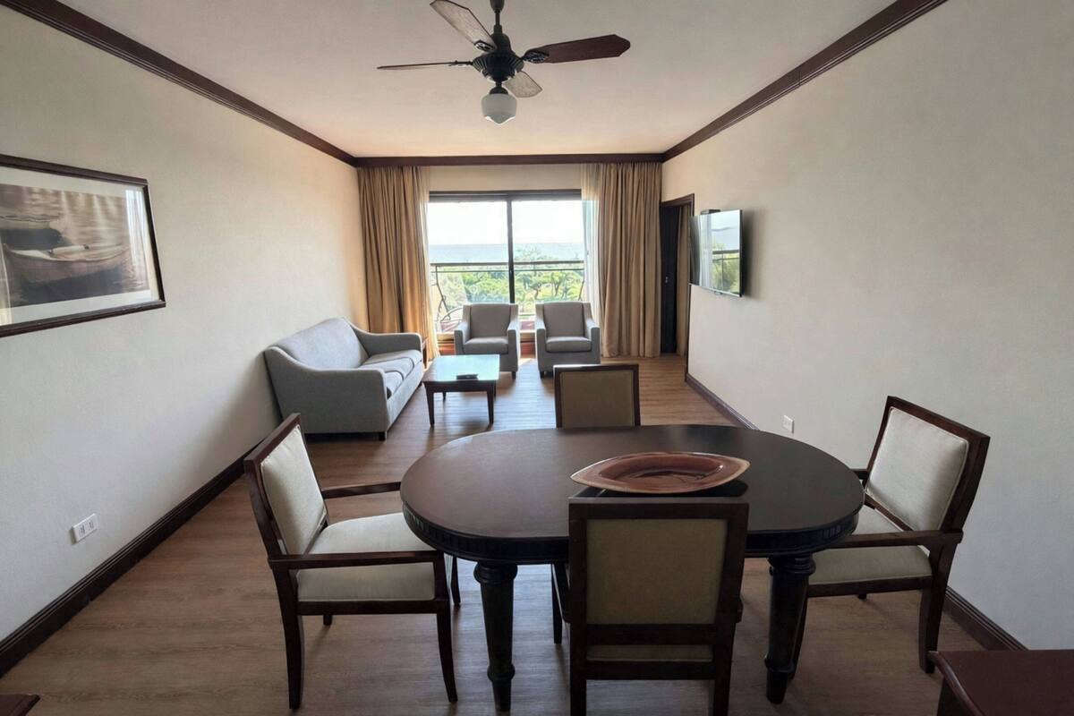 Un comedor con una mesa redonda y sillas, un ventilador de techo y vistas al exterior a través de amplias ventanas.
