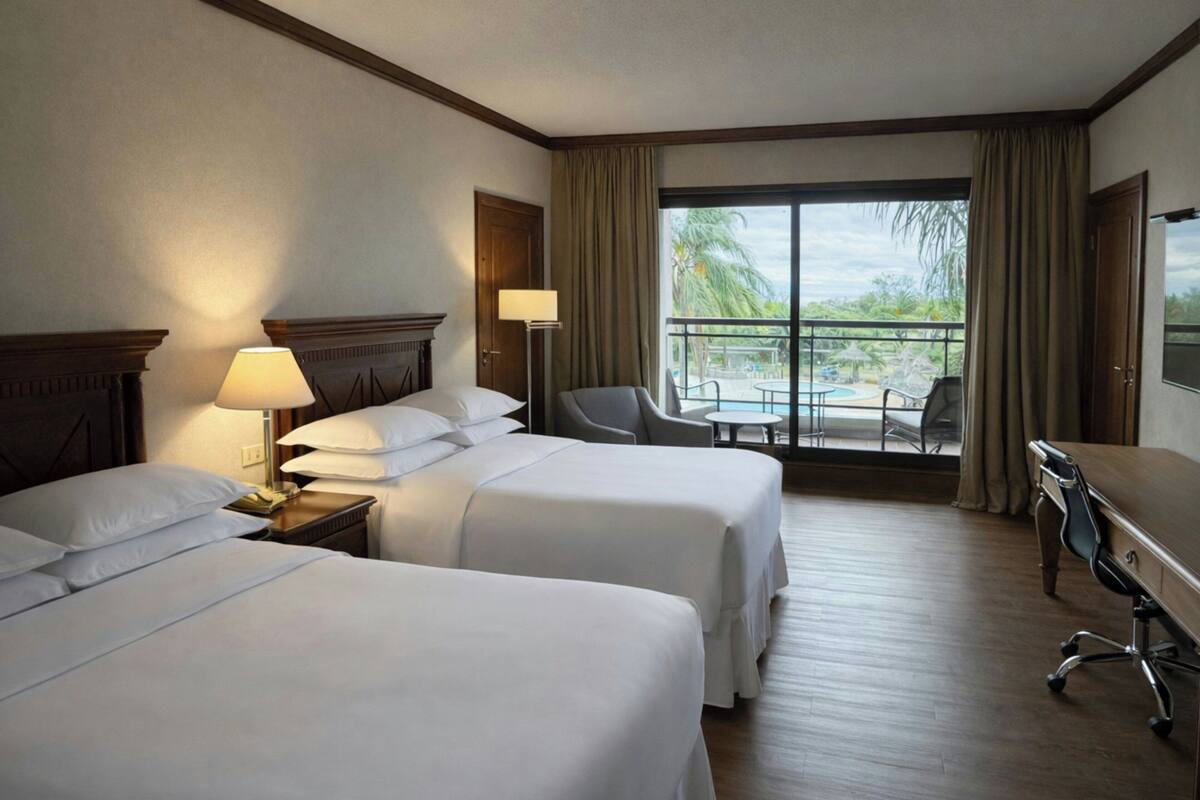 Habitación de hotel con dos camas, un escritorio, una silla y un balcón con vista a la piscina.