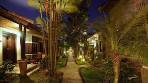 A pathway flanked by palm trees and greenery, leading to a building with a thatched roof and wooden doors. - Putu Bali Villa and Spa (Seminyak)