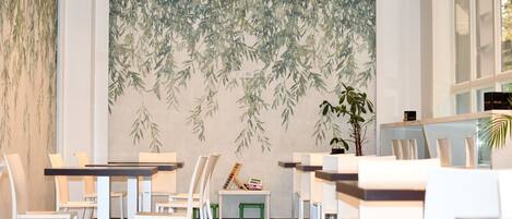 Un comedor moderno con sillas y mesas blancas, un mural de hojas de bambú y una planta en maceta.