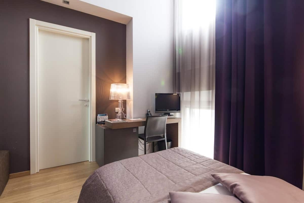 Una habitación de hotel con una cama, un escritorio con una computadora, una lámpara y una puerta.