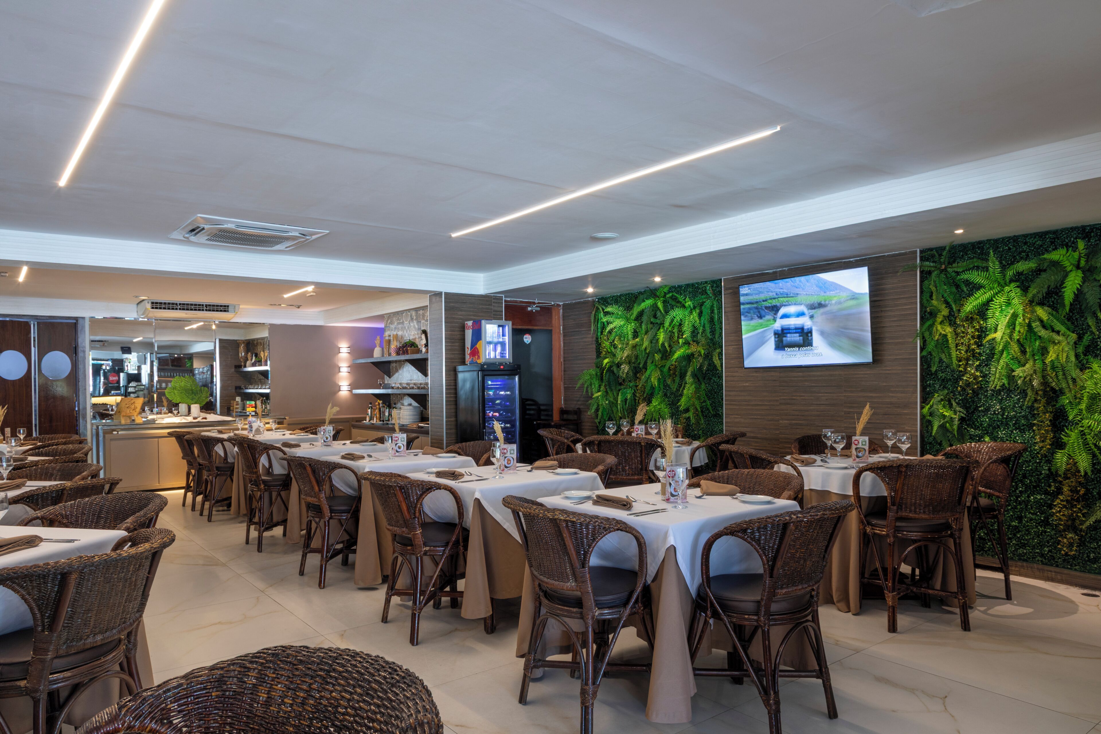Un restaurant avec plusieurs tables et des chaises en osier, un espace cuisine et un grand écran sur lequel est affichée une image de voiture.