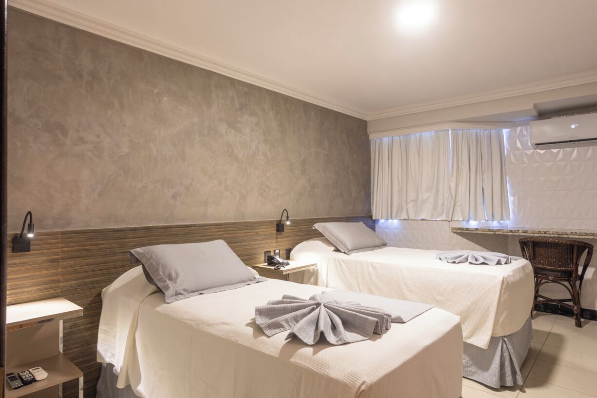 A hotel room with two beds, a bedside table with a lamp, a chair, and an air conditioning unit.