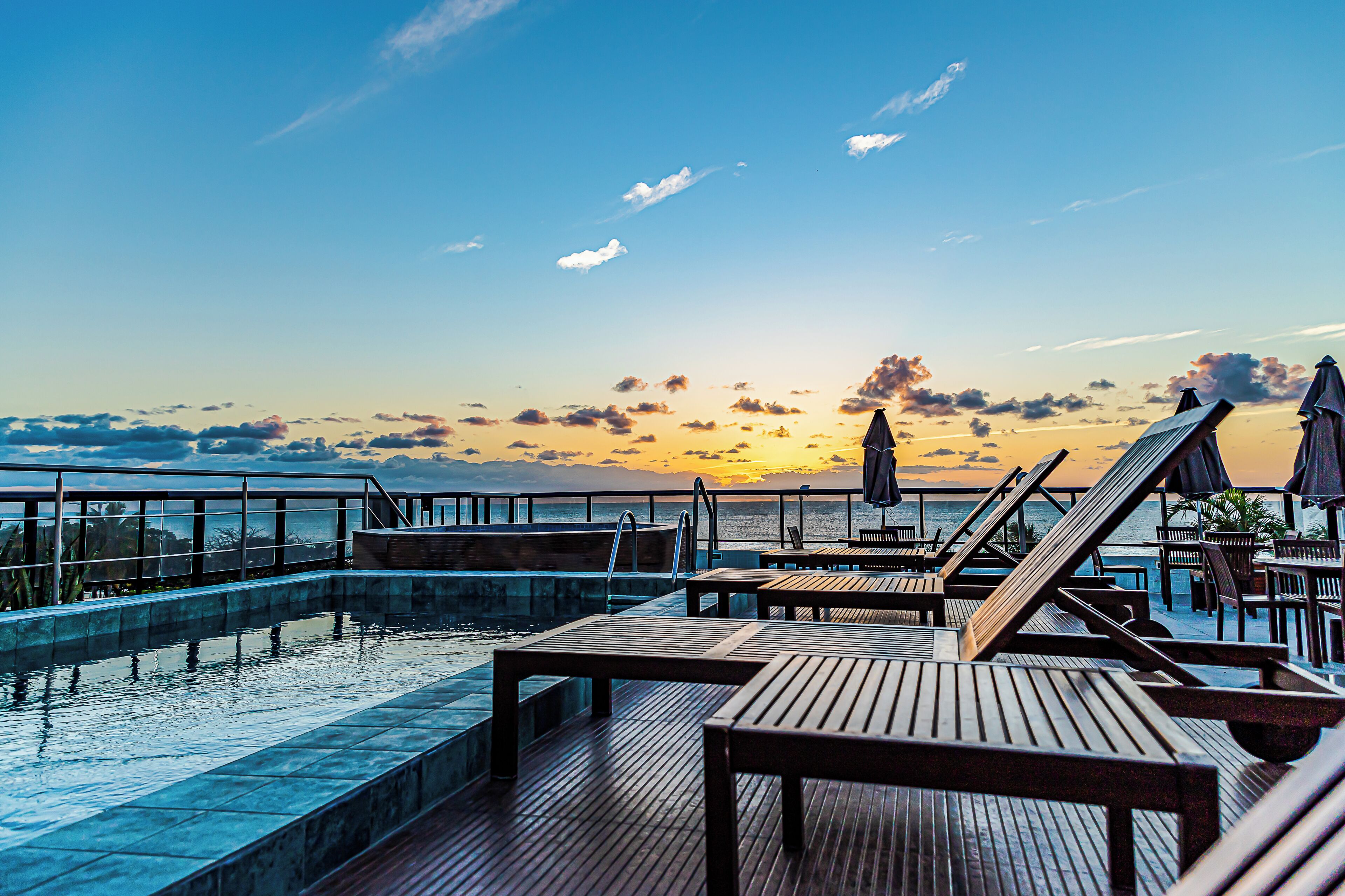 Zona de piscina con sillones y mesas, vista al atardecer sobre el agua, y una persona caminando a lo lejos.