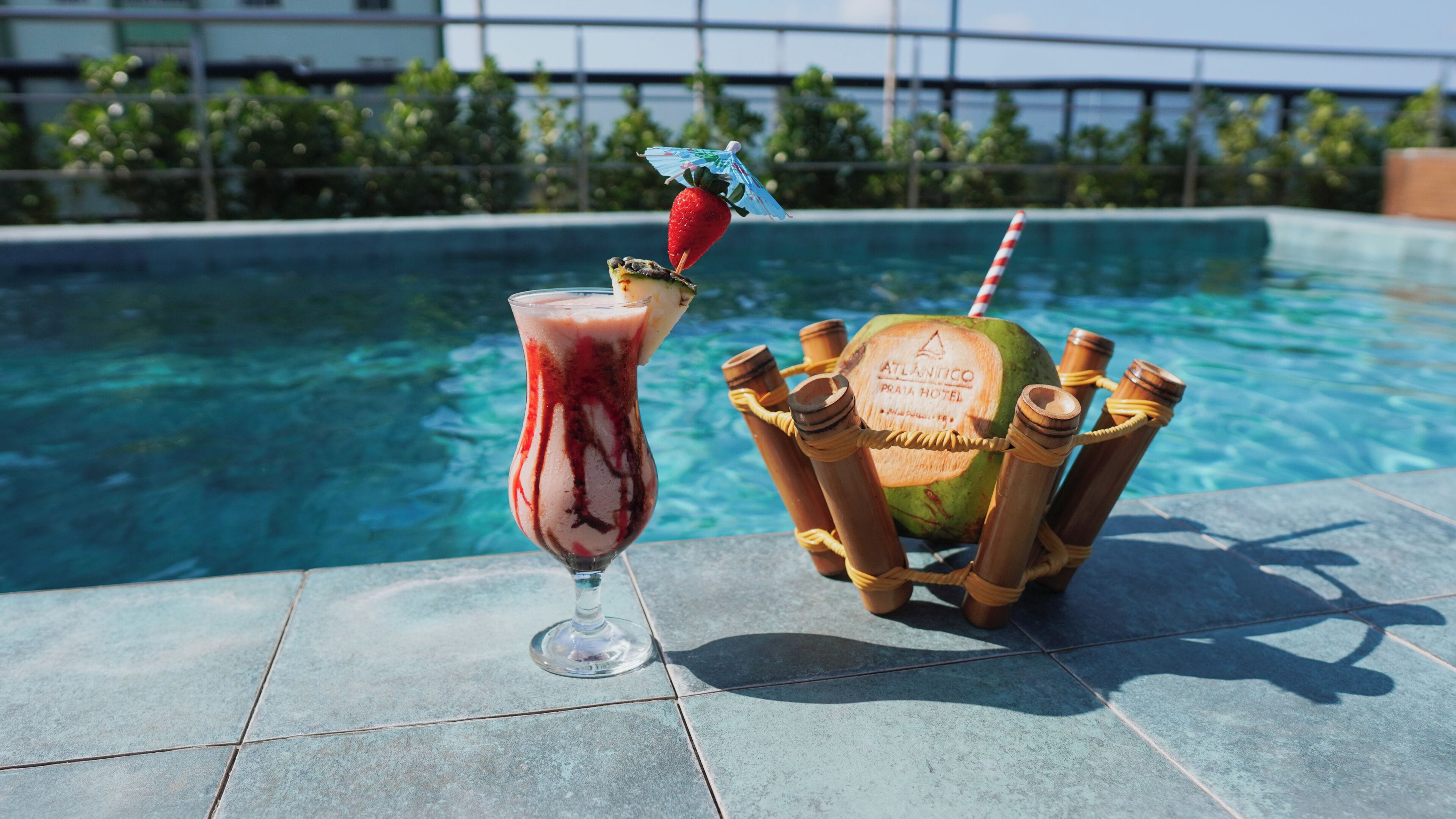 A poolside setting with a drink in a glass and a coconut with a straw.