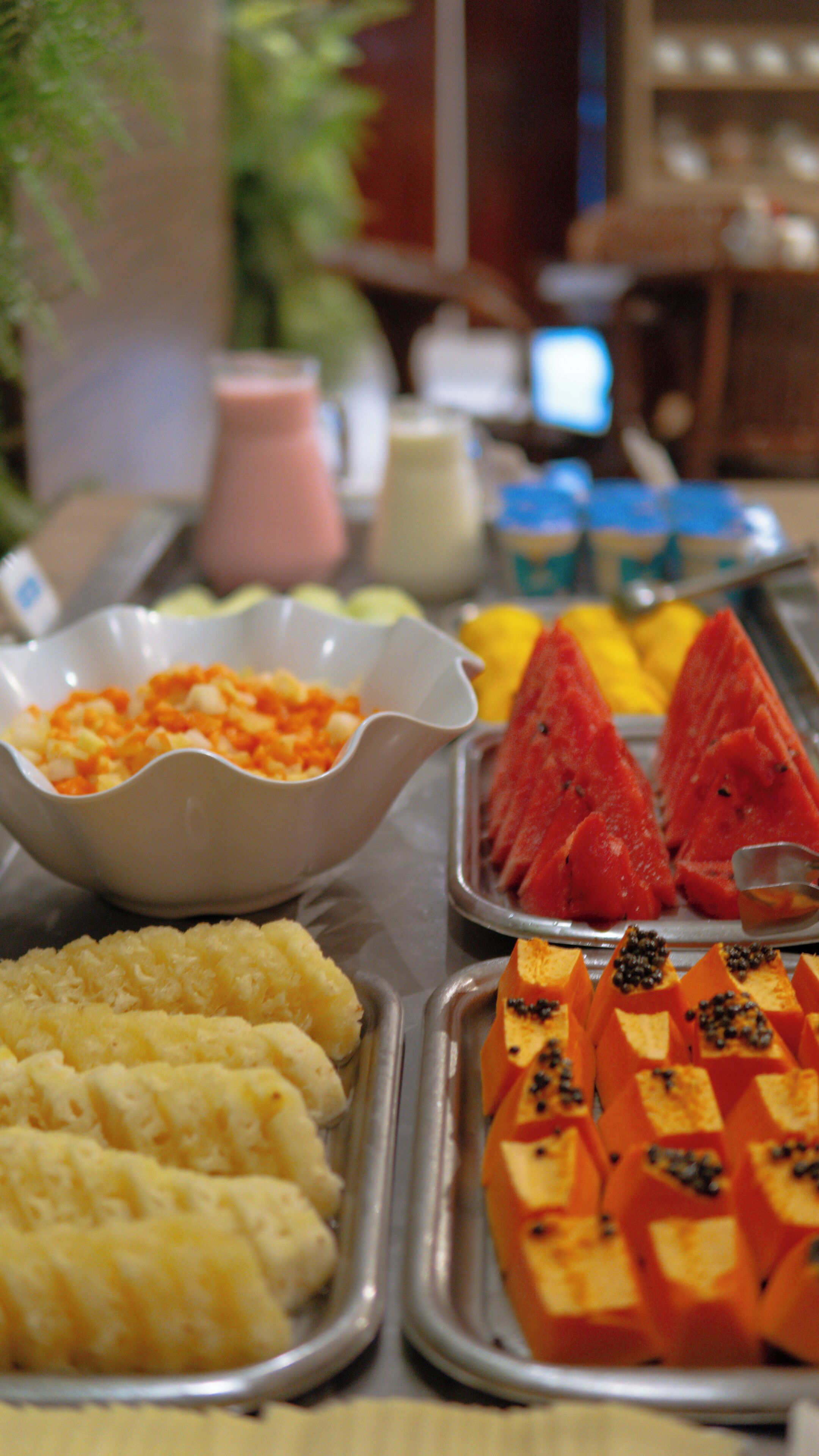 Uma mesa de buffet com diversos alimentos, incluindo uma tigela de legumes picados, uma bandeja de melancia em fatias e um prato com uma substância amarelada.