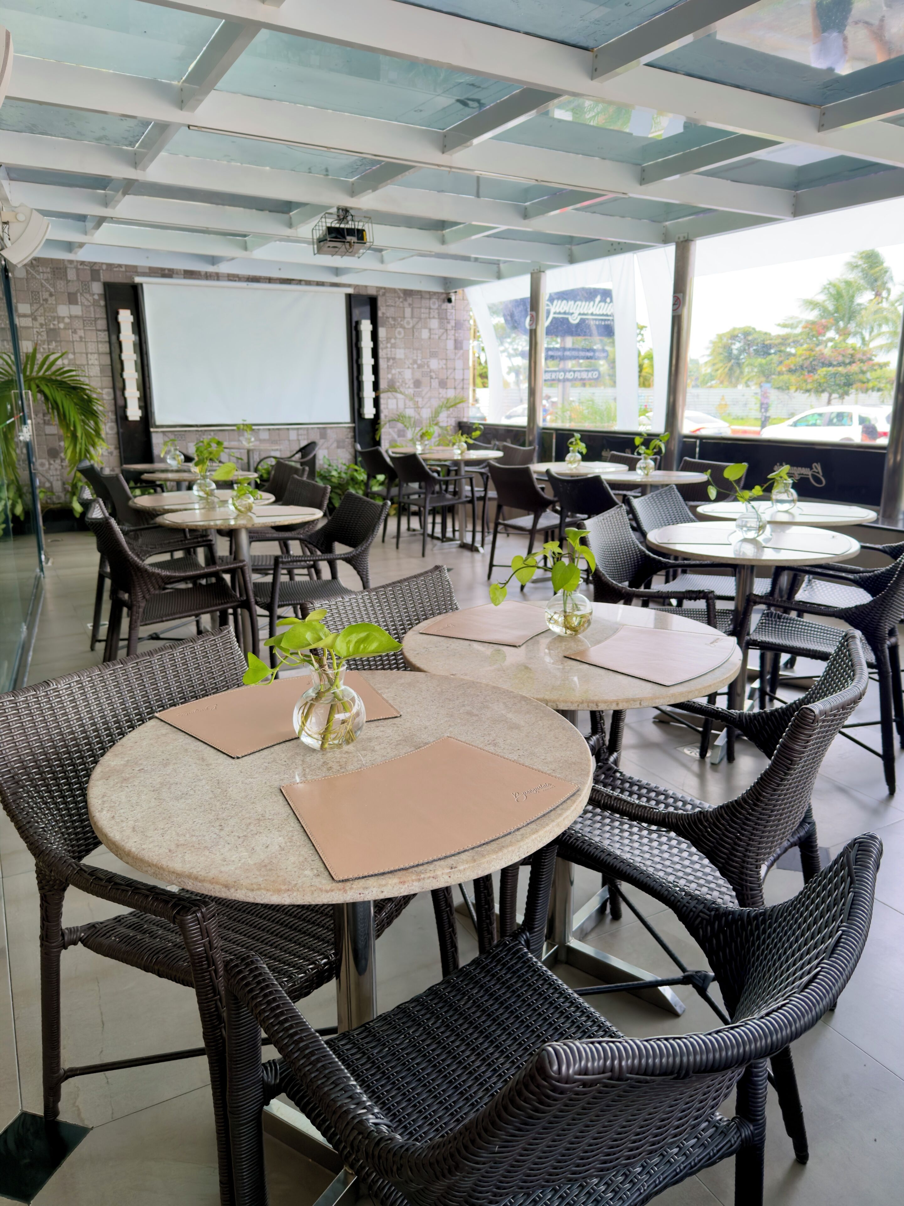 A restaurant with round tables and wicker chairs, a projector screen, and a view of the outside.