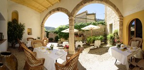 An outdoor dining area with tables and chairs, surrounded by stone walls and arches, featuring potted plants and umbrellas. - Finca Son Palou (Bunyola)
