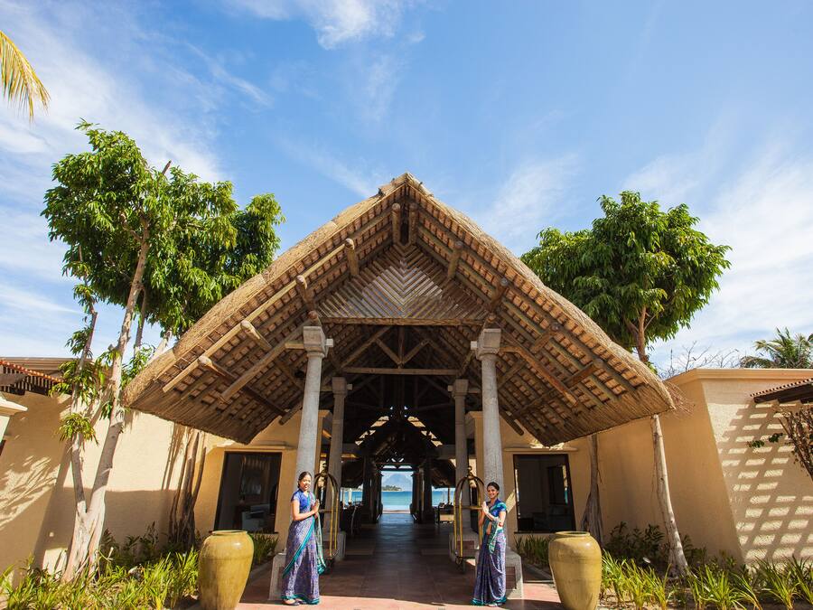 A covered entrance with a thatched roof, palm trees, and a view of the sea.