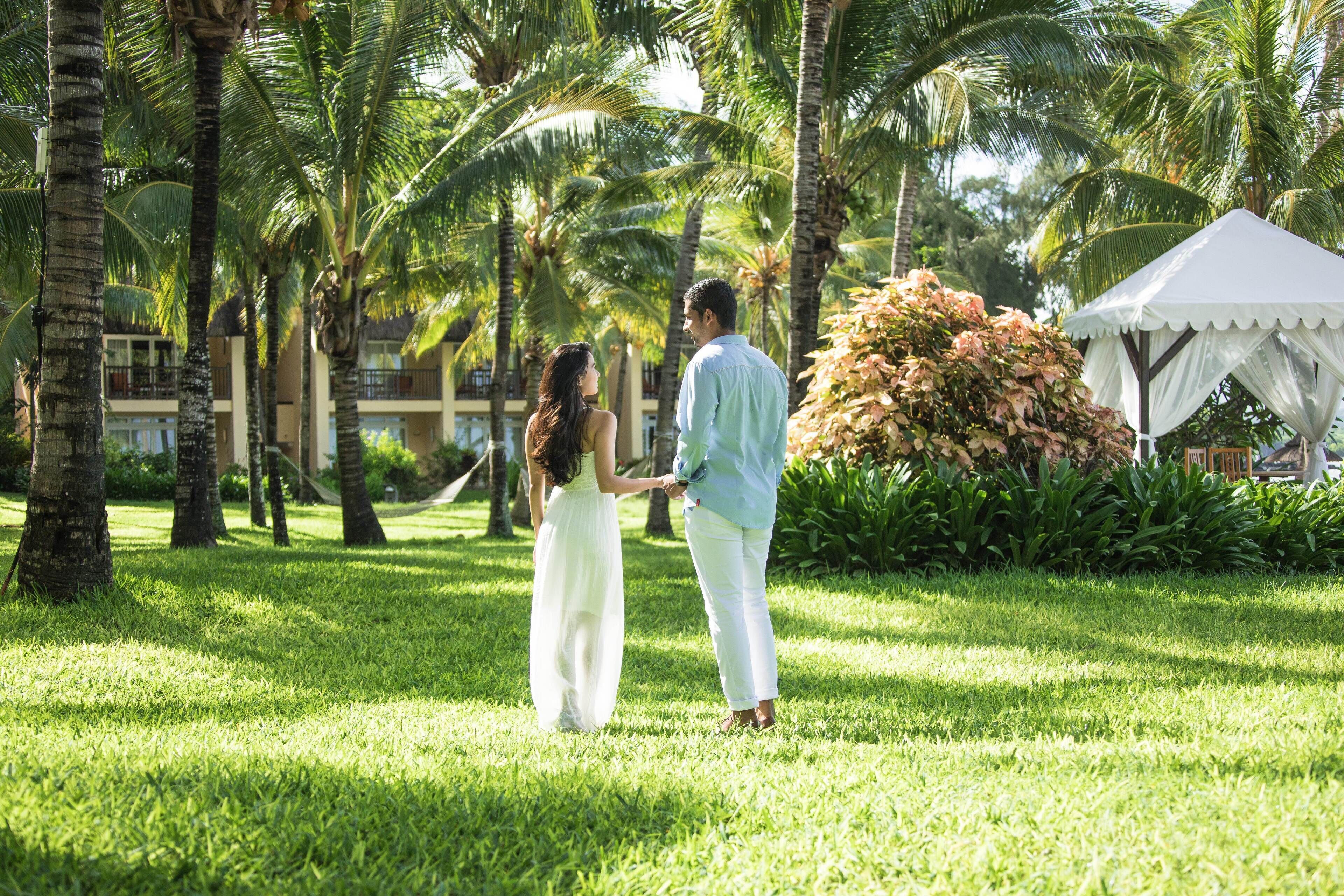 A couple holding hands in a lush, green outdoor area with palm trees and a white tent.