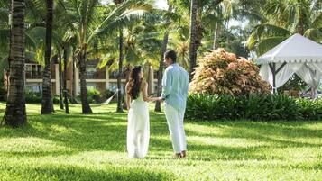 A couple holding hands in a lush, green outdoor area with palm trees and a white tent.