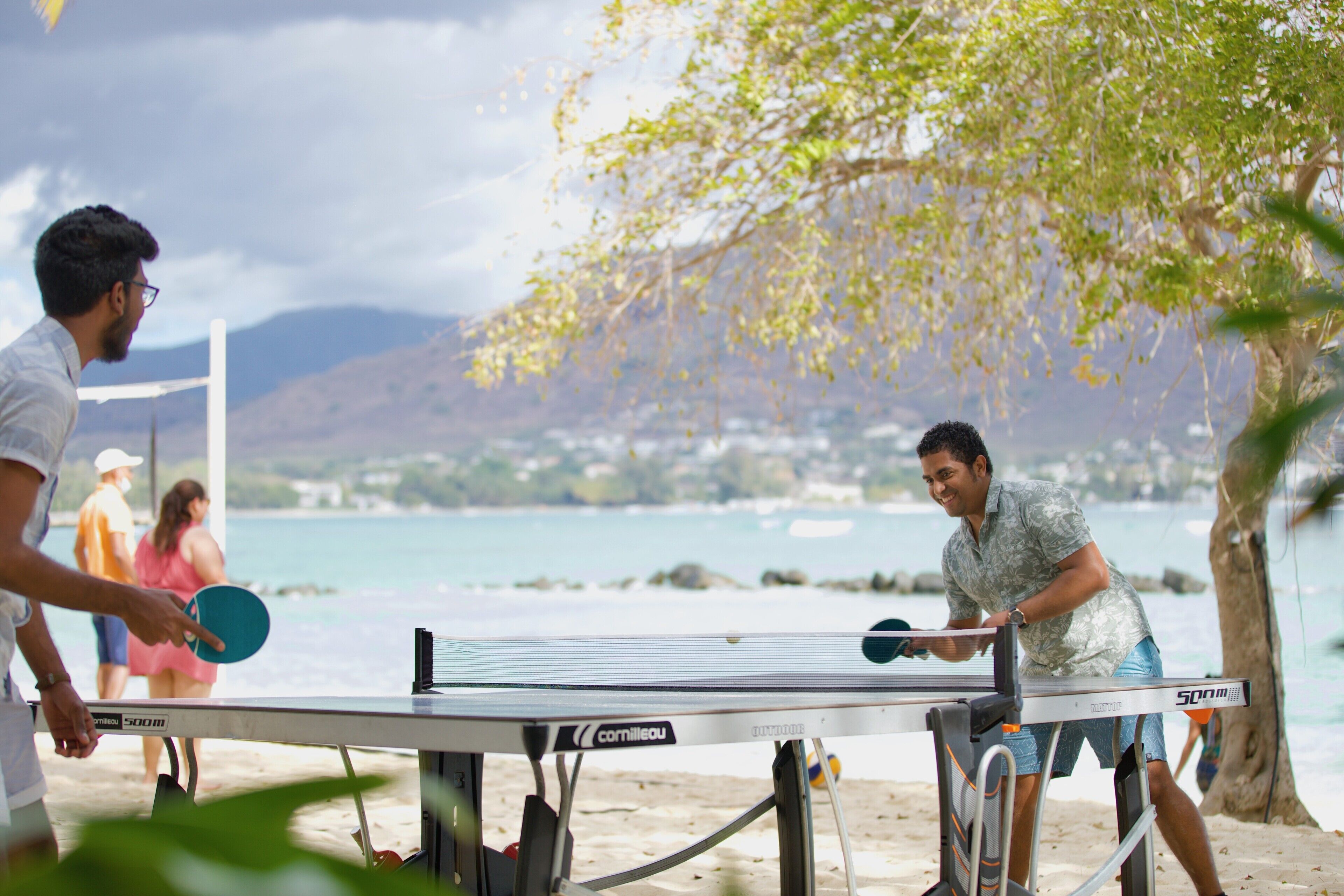 A table tennis table with two players engaged in a match, set against a beach backdrop with a view of the sea and distant hills.