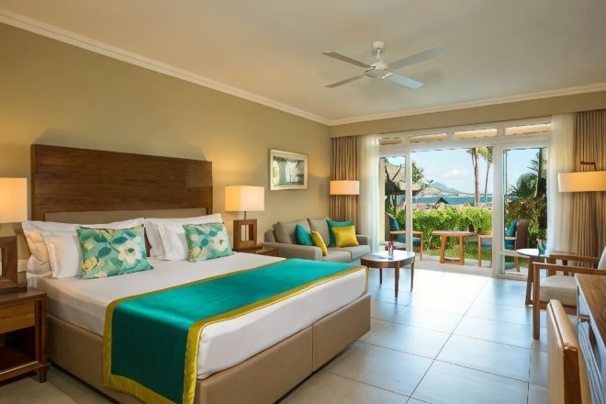 Ein Hotelzimmer mit einem großen Bett, einer Sitzecke mit Sofa und Sesseln, einem Balkon mit Blick ins Freie und einem Deckenventilator.