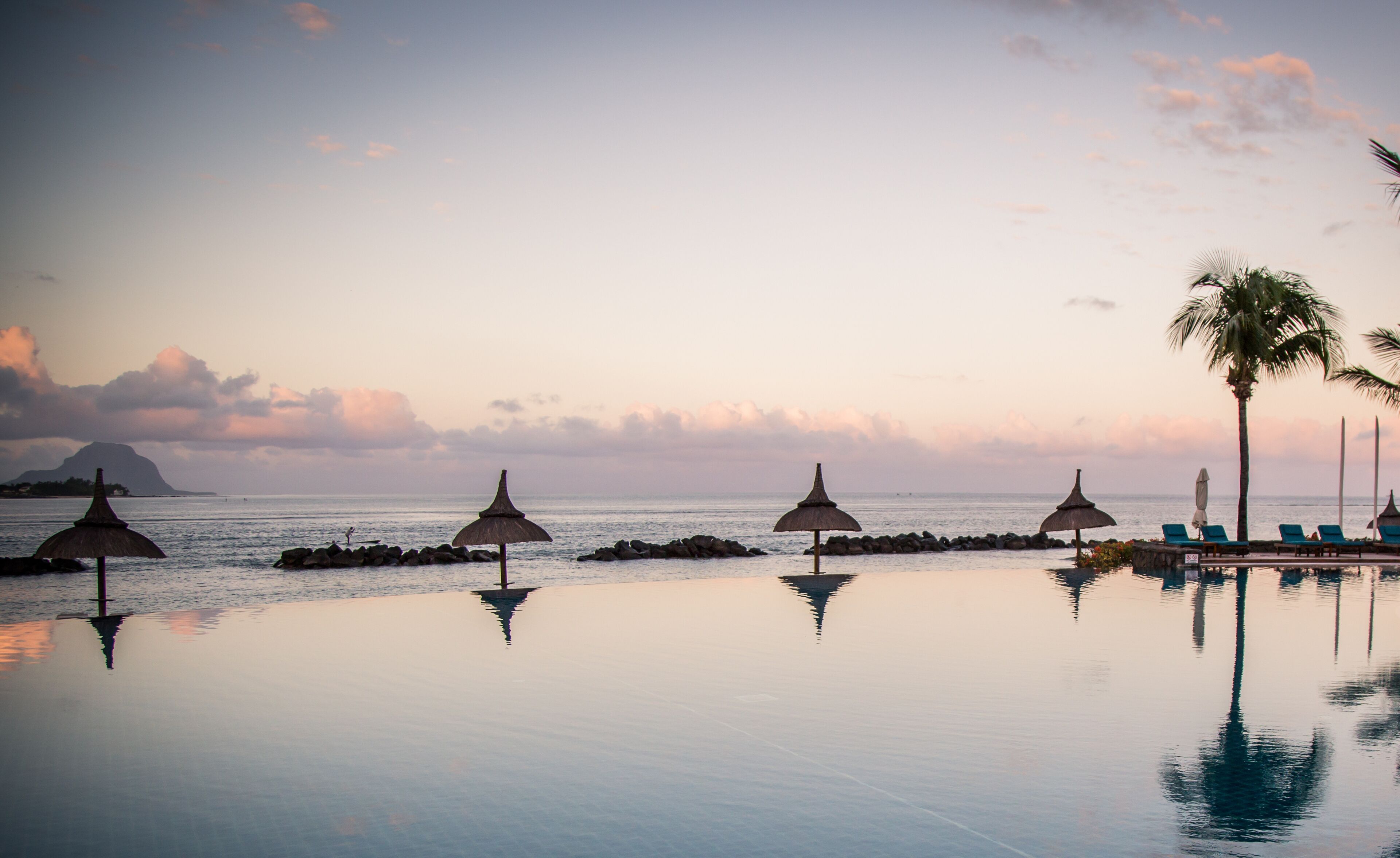 Una piscina infinita tranquila con sombrillas de paja, una palmera y vistas al océano al atardecer.