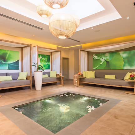 A modern spa room with two sofas, a central water feature, and large green leaf wall art.