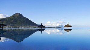 Une scène de plage paisible, avec une montagne en arrière-plan et trois parasols à toits de chaume dont le reflet se mire dans l’eau.