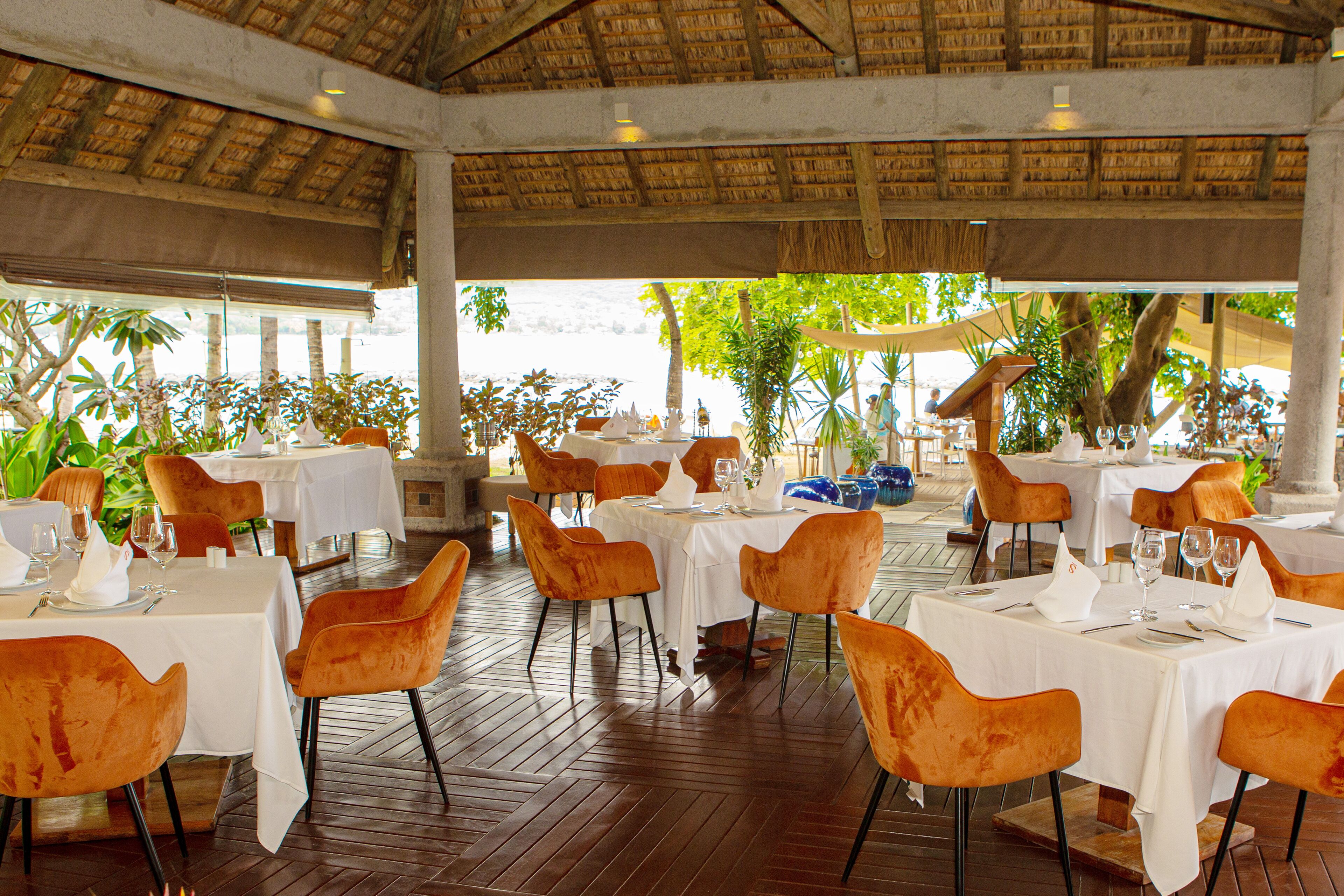 A restaurant with tables and chairs, a thatched roof, and a view of the sea.
