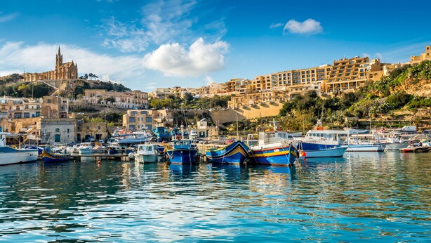 Un puerto con barcos amarrados, una iglesia histórica en una colina y edificios residenciales.