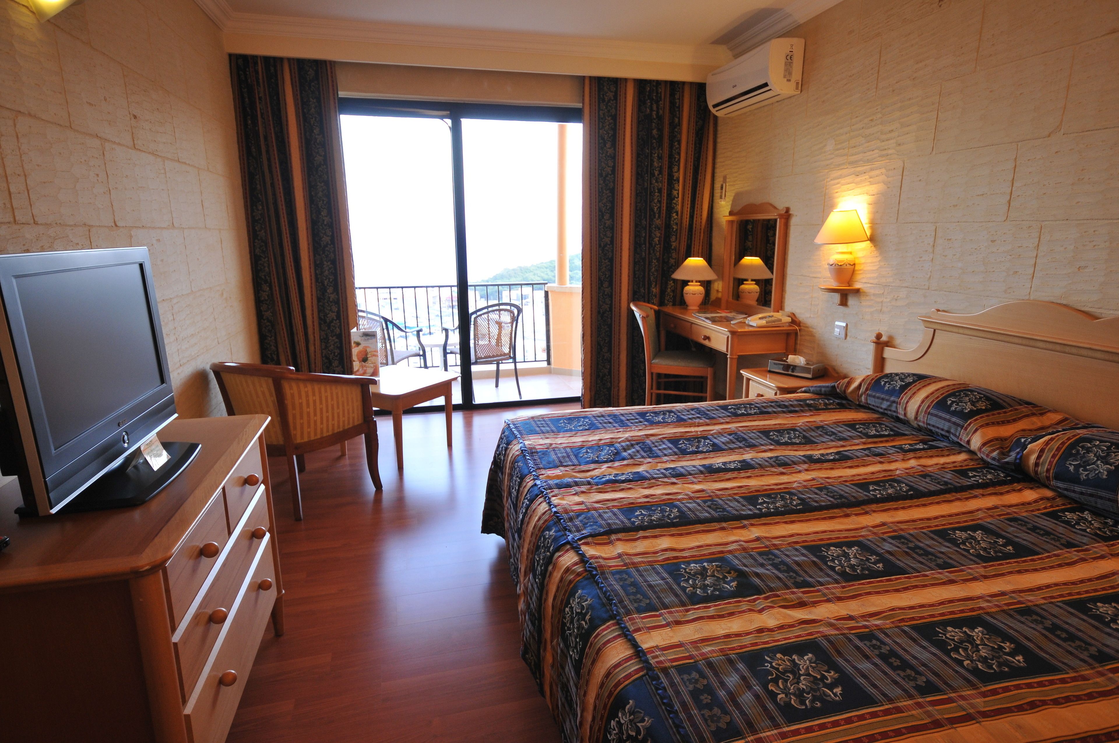 A hotel room with a bed, a desk, a chair, a television, and a balcony with a view.