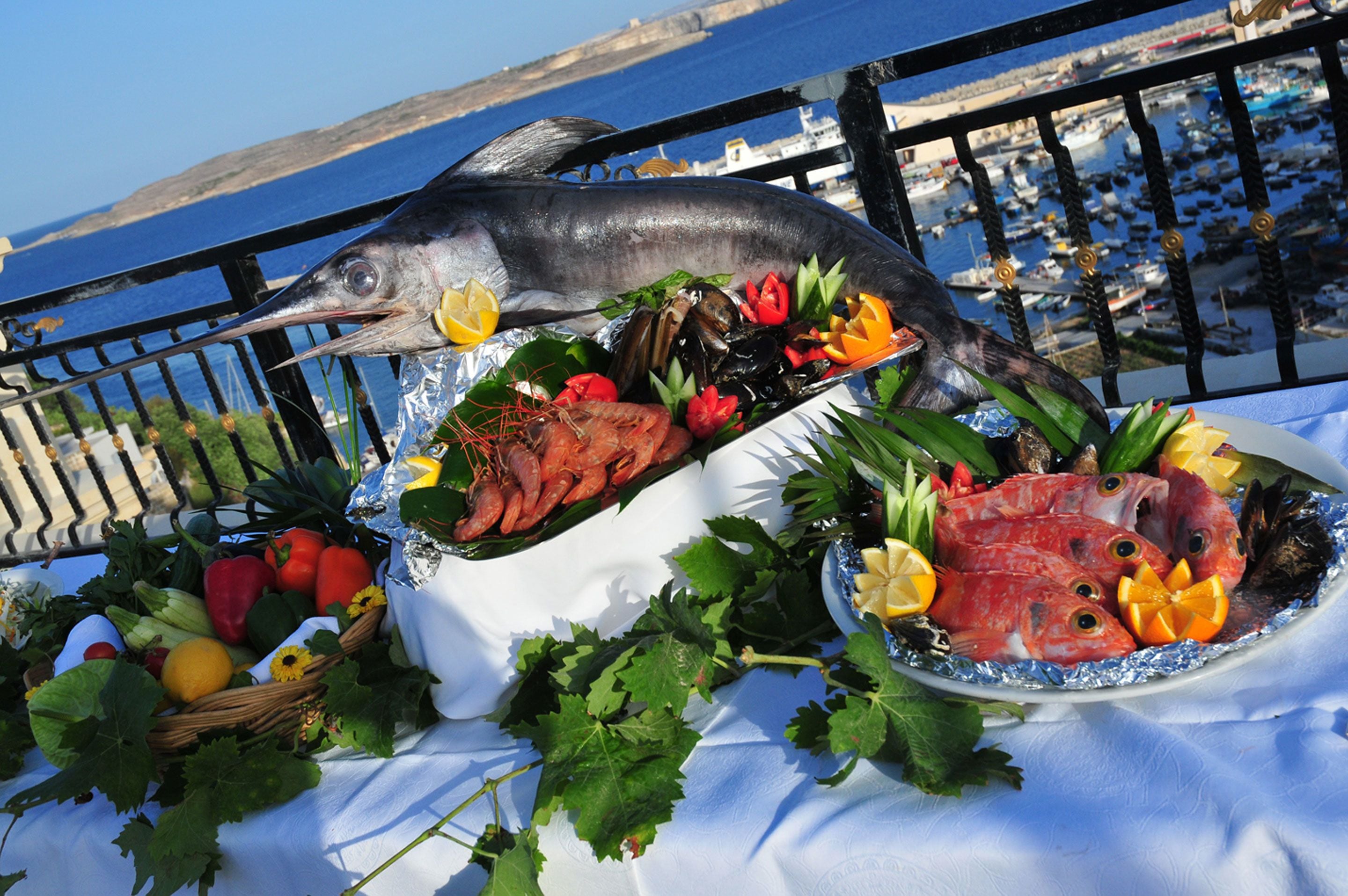 Una bandeja de mariscos con varios pescados y verduras sobre una mesa, con vista al mar y embarcaciones al fondo.