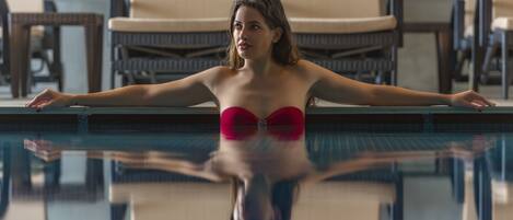 Una persona en traje de baño rojo al borde de una piscina, con sillones y mesas al fondo.