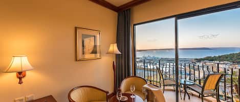 A hotel room with a balcony, two chairs, a table, a lamp, a desk, and a framed picture.