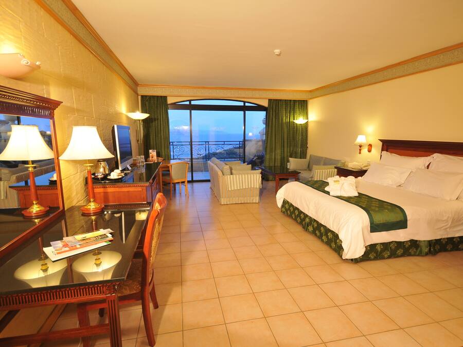 A hotel room with a large bed, a desk with a chair, a small table, and a view of the sea through a large window.
