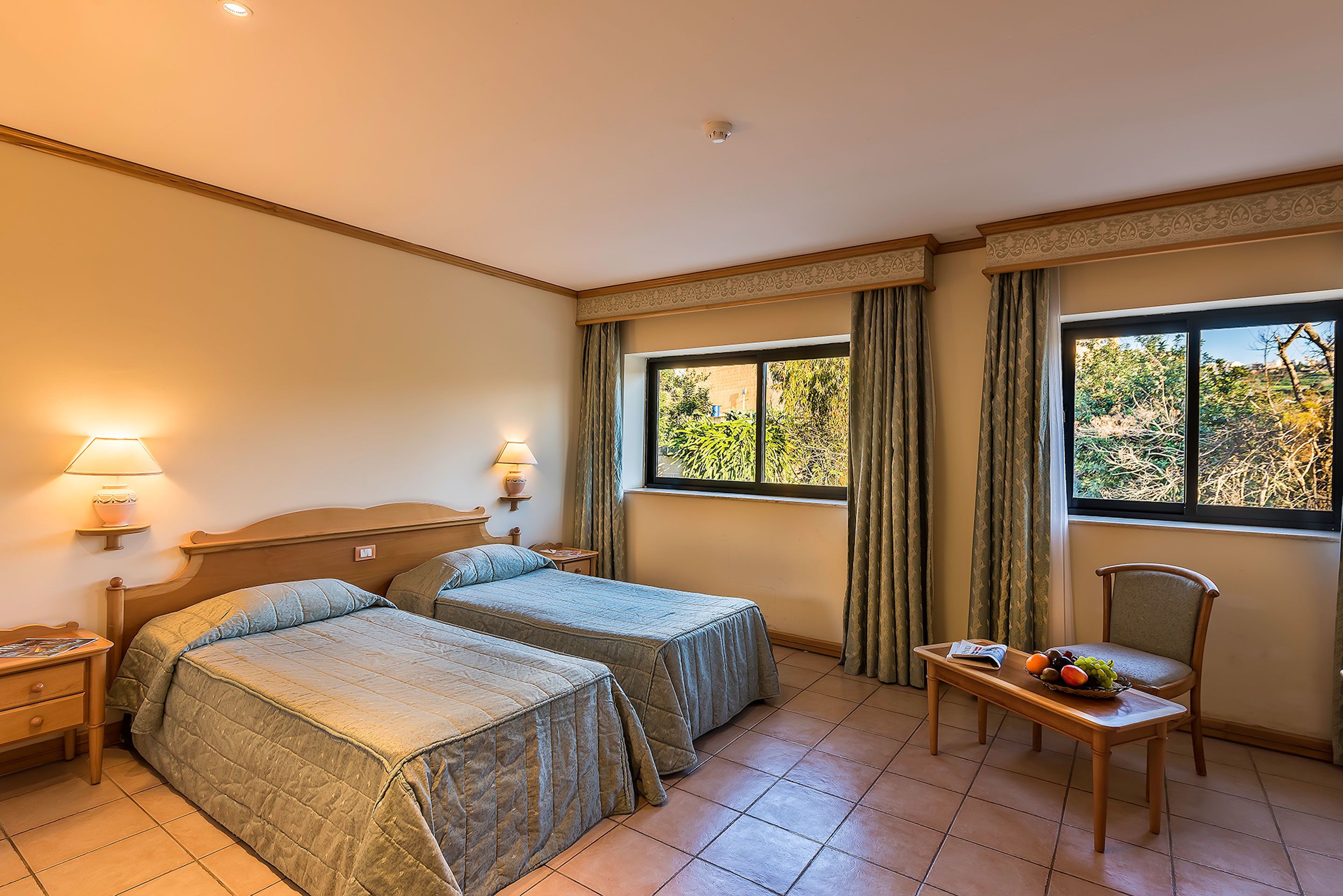 Una habitación de hotel con dos camas, una mesita con fruta, una silla y un ventanal con cortinas.