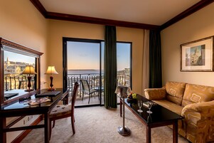 Habitación de hotel con balcón, zona de comedor, sofá y una mesa con una copa de vino.