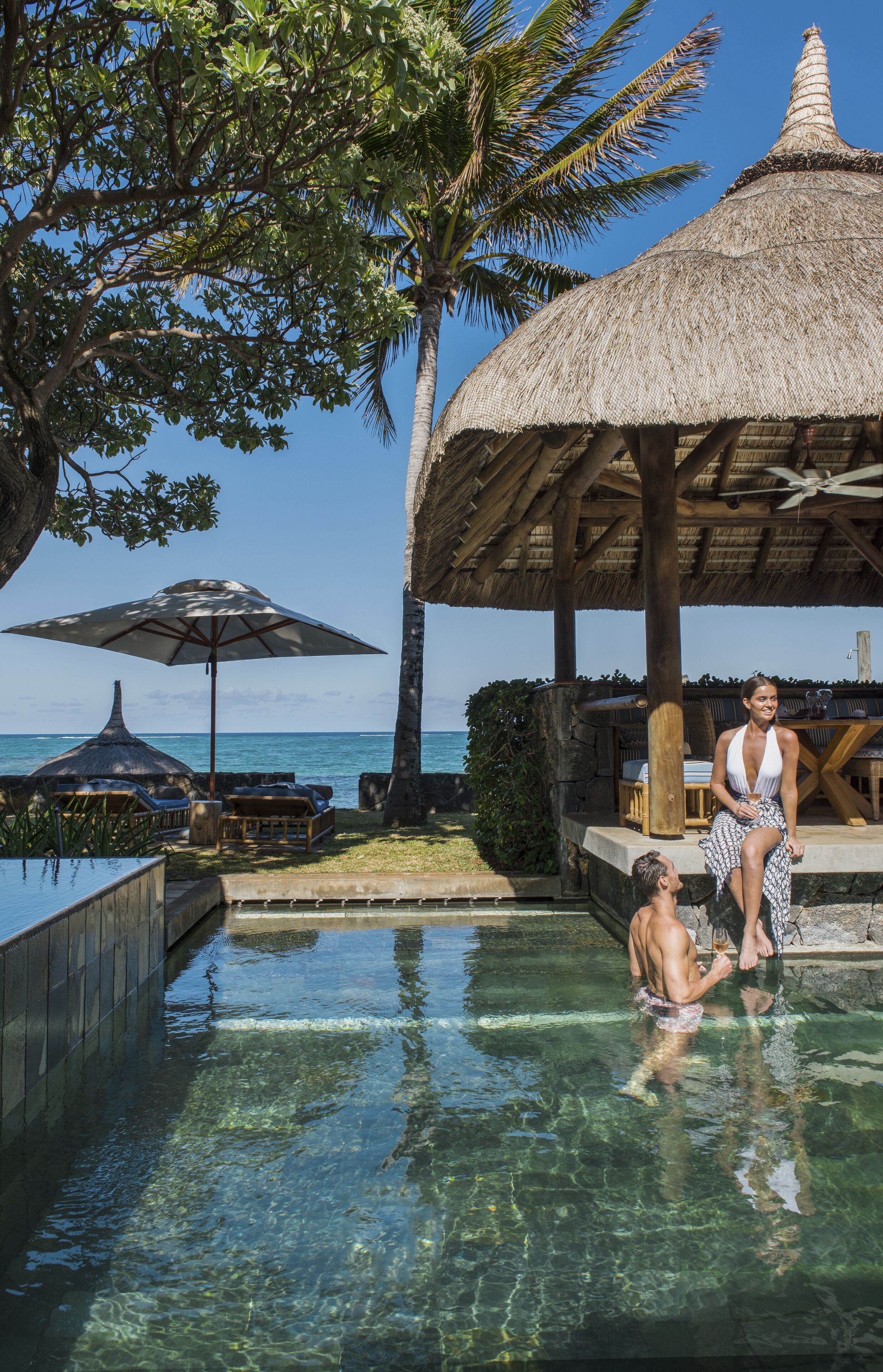 A pool area with clear water, a thatched-roof structure, and a view of the ocean.