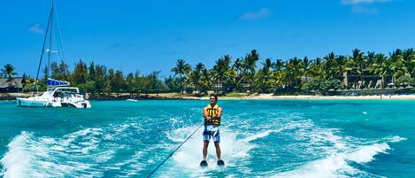A person wakeboarding on turquoise waters with a sailboat in the background and a beach with palm trees.