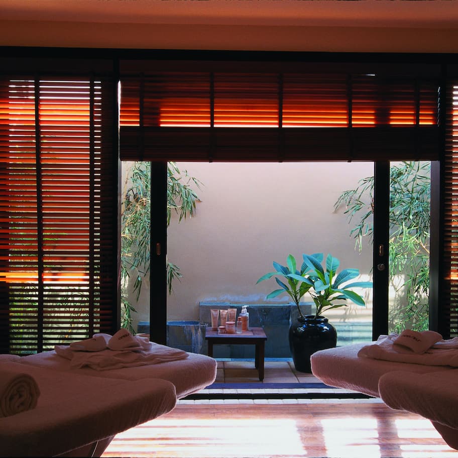 A spa room with two massage tables, a small table with toiletries, and a view of a garden through a sliding door.