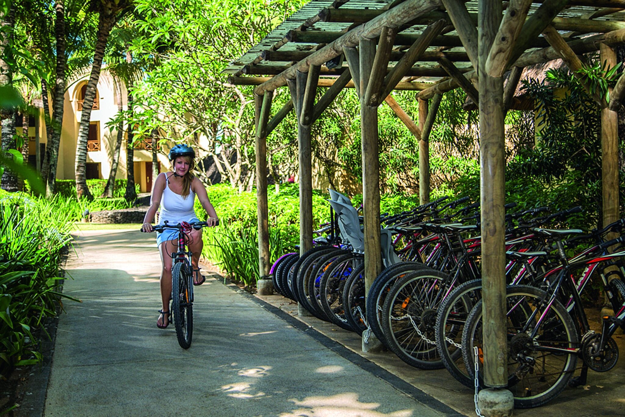 A woman riding a bicycle on a pathway with a row of bicycles parked under a wooden pergola.