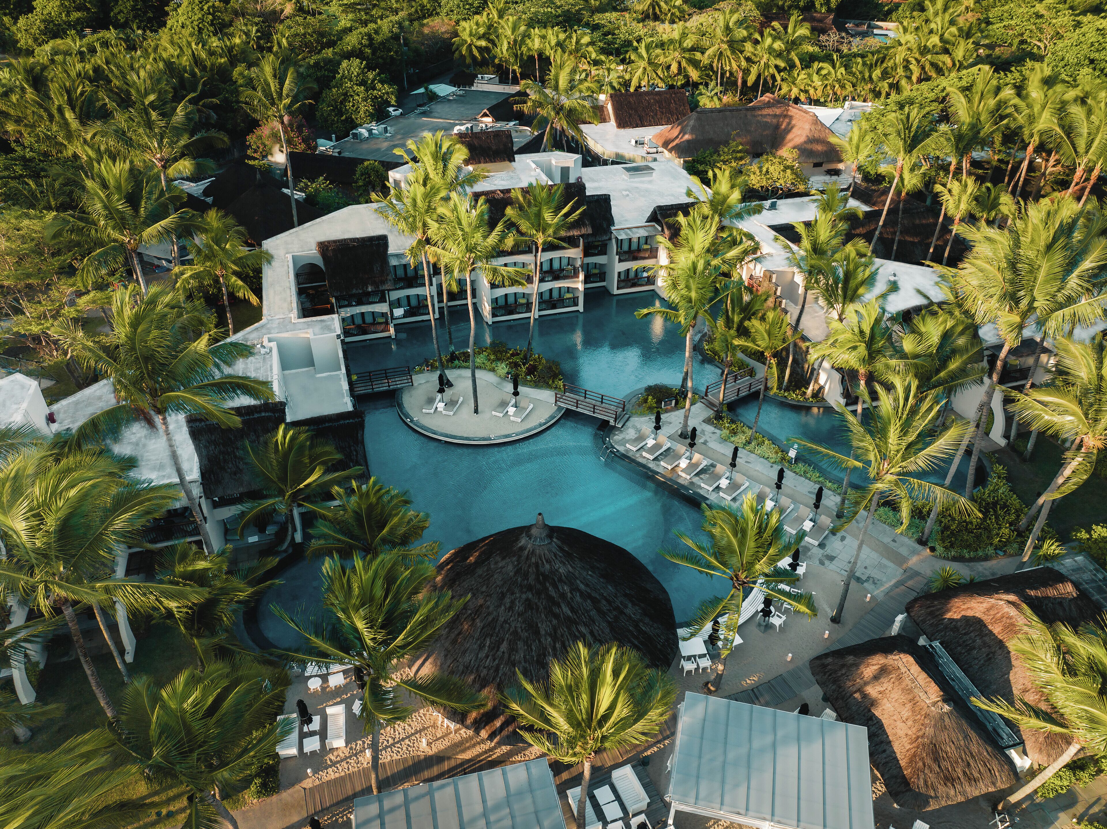 An aerial view of a resort with a central pool, surrounded by palm trees and thatched-roof buildings.