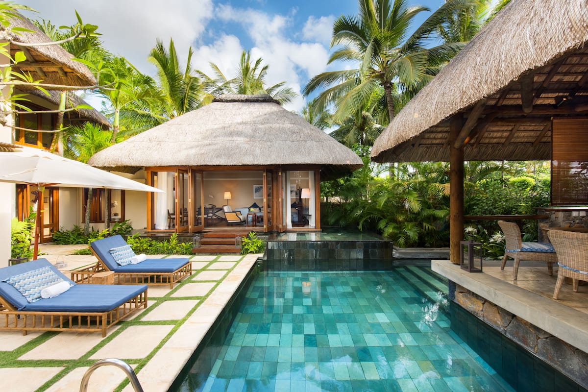 Un área de piscina con sillones de descanso, una estructura con techo de paja y follaje tropical.