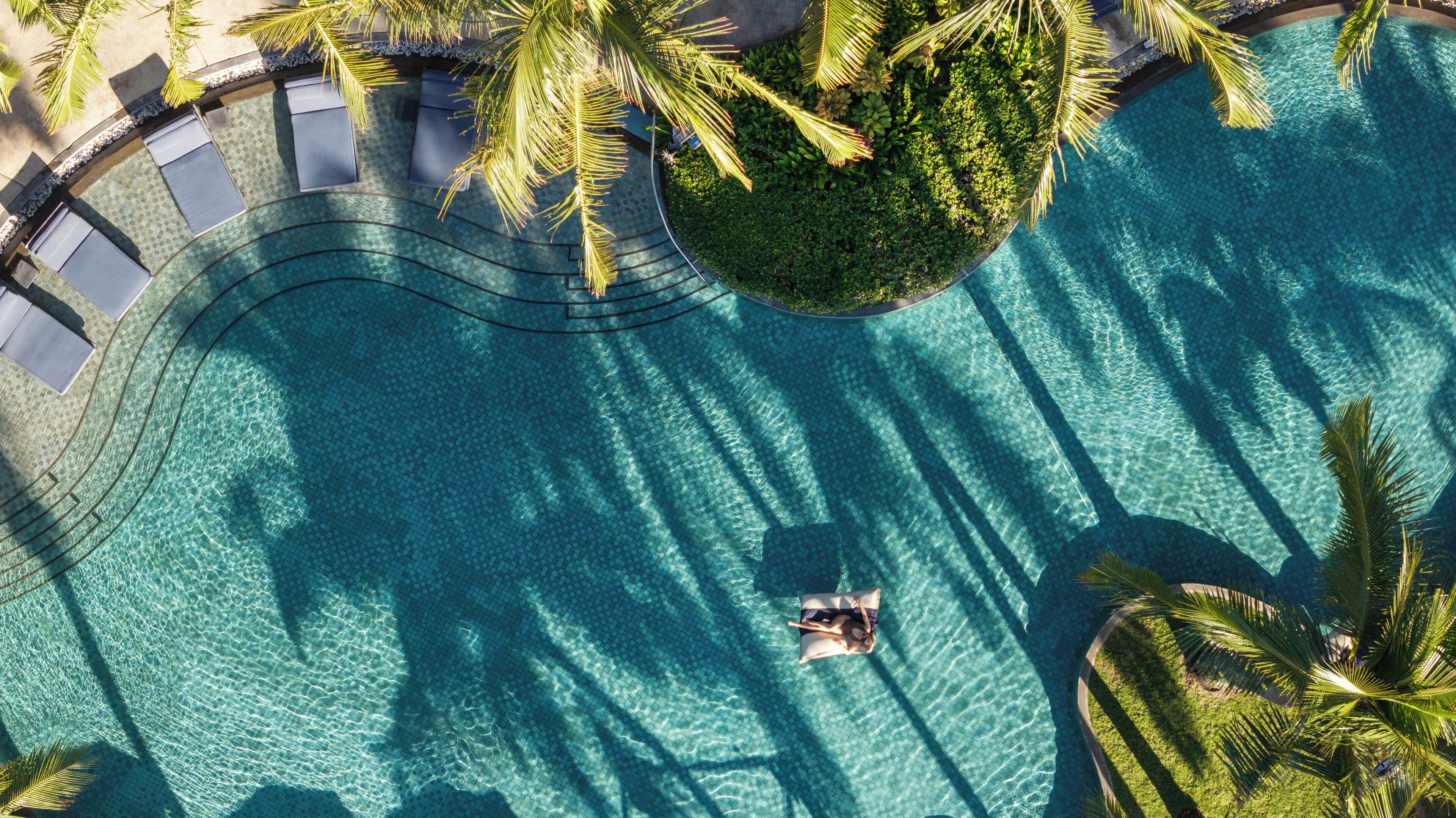 Vista aérea de una piscina con agua azul cristalina, rodeada de palmeras y un sendero curvo.