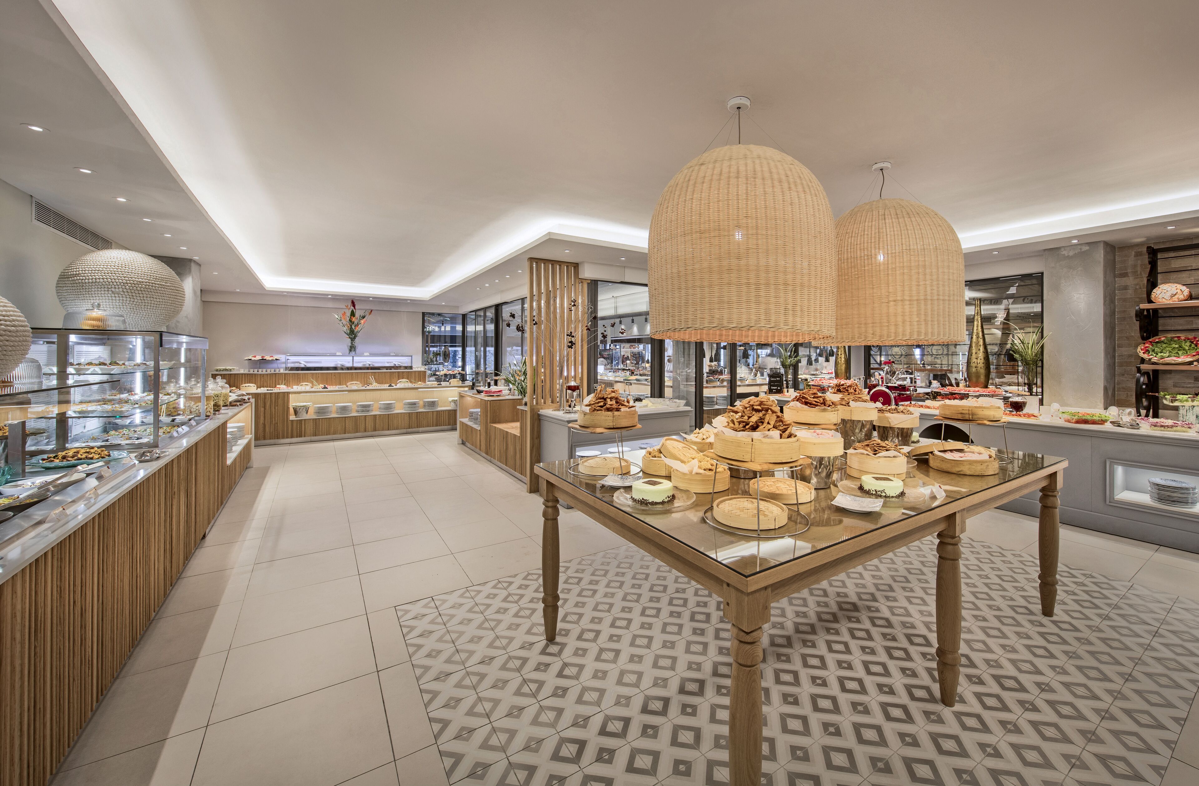 A modern buffet area with a variety of desserts displayed on a glass table, surrounded by wooden paneling and a patterned floor.