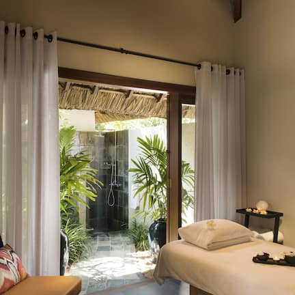 A room with a bed, a massage table, a small table with a bowl of flowers, and a view of a tropical garden through a glass door.