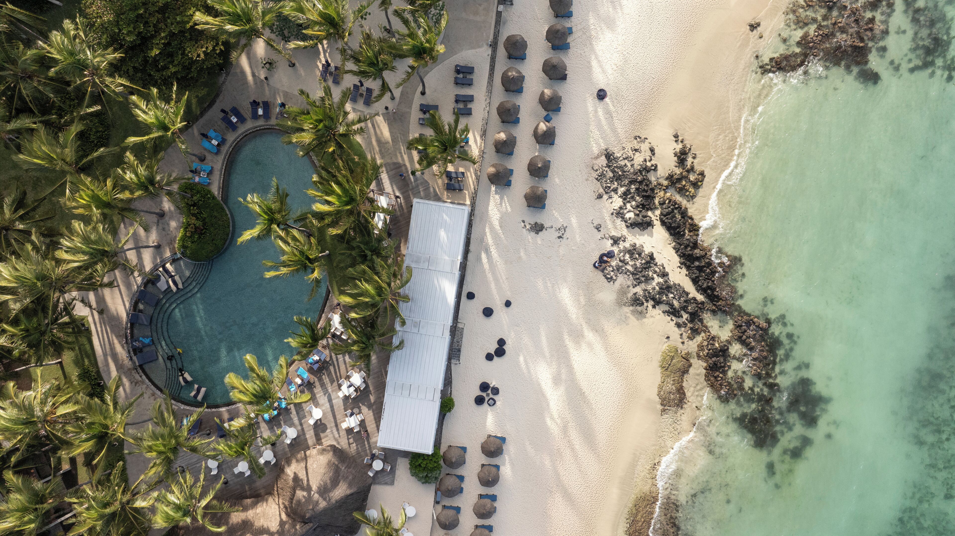 An aerial view of a beach resort with a swimming pool, lounge chairs, umbrellas, and palm trees.