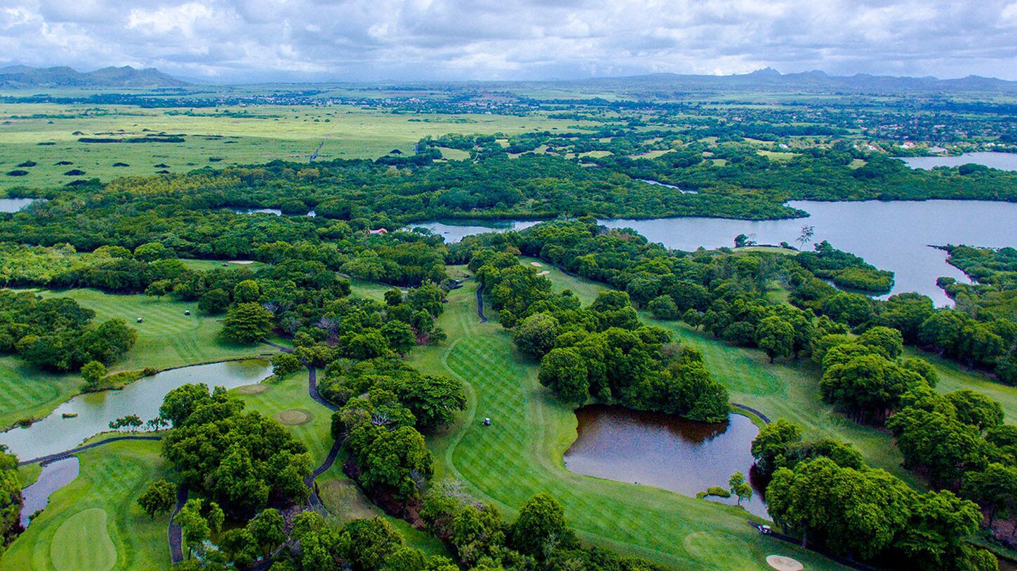 An aerial view of a golf course with multiple water hazards, surrounded by lush greenery and distant mountains.