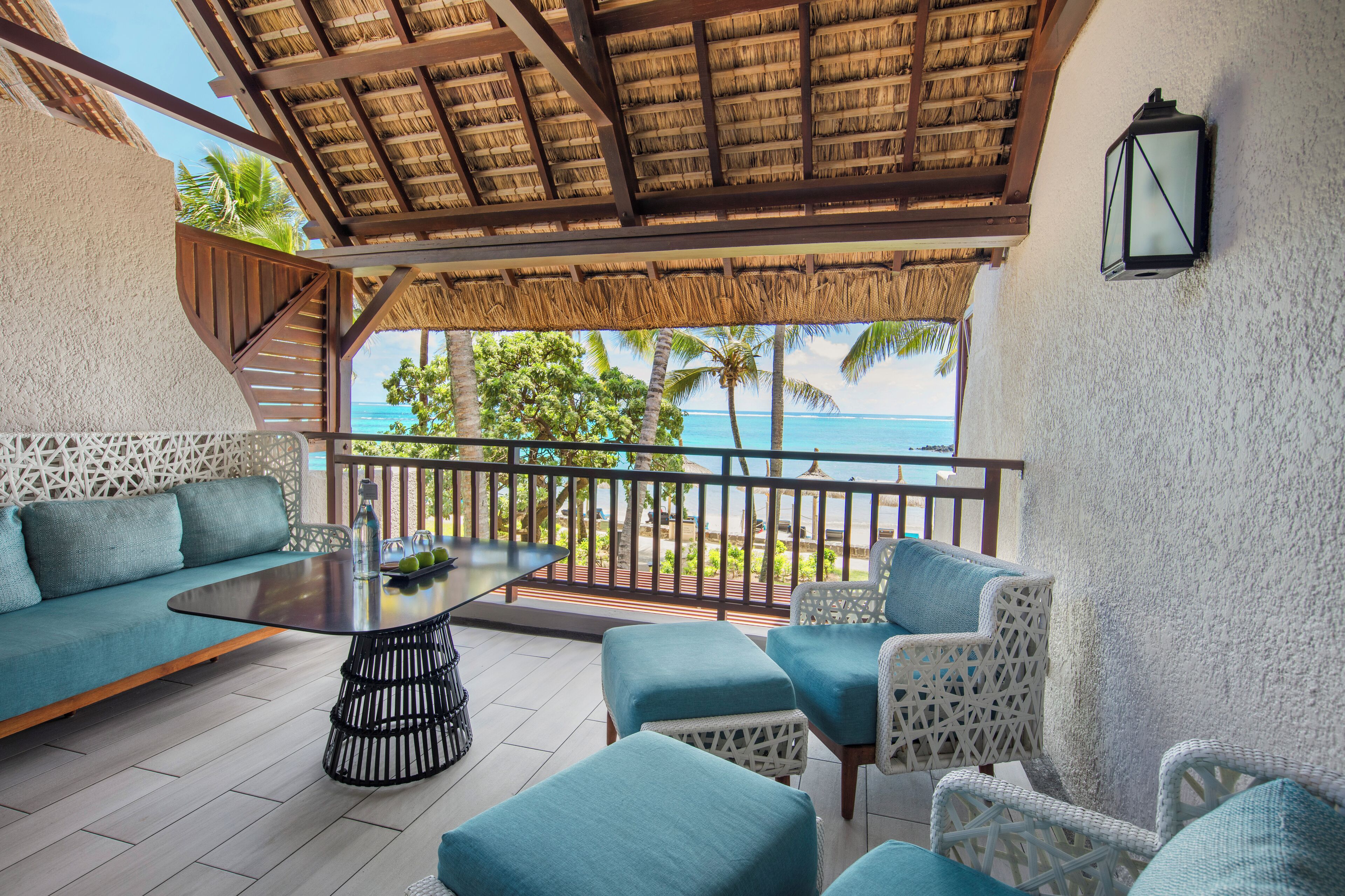 A balcony with a thatched roof, a sofa, and wicker chairs, overlooking a beach and palm trees.