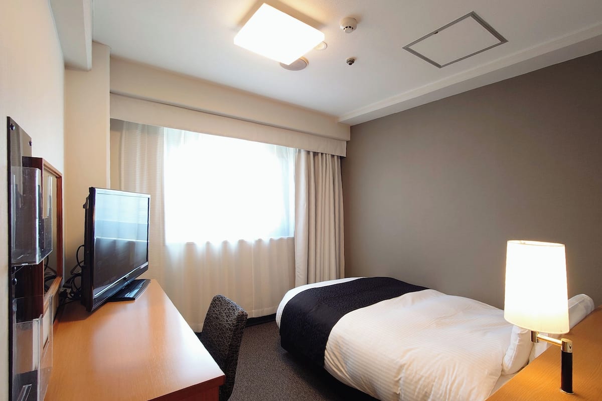 Ein Hotelzimmer mit einem Bett, einem Schreibtisch mit Fernseher, einer Lampe und einem Fenster mit Vorhängen.