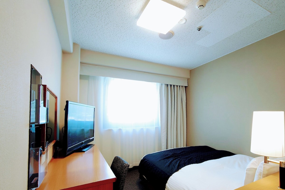 Ein Hotelzimmer mit einem Bett, einem Schreibtisch mit Fernseher, einer Lampe und einem Fenster mit Vorhängen.