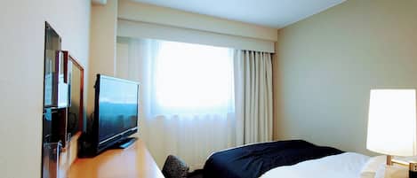 A hotel room with a bed, a desk with a television, a lamp, and a window with curtains.
