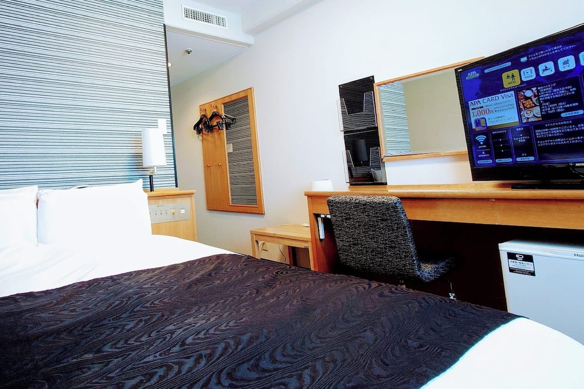 Ett hotellrum med en säng, ett skrivbord, en stol, en tv och ett fönster med persienner.