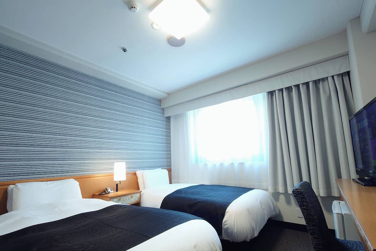 Ein Hotelzimmer mit zwei Betten, einem Schreibtisch, einem Stuhl, einem Fernseher und einem Fenster mit Vorhängen.