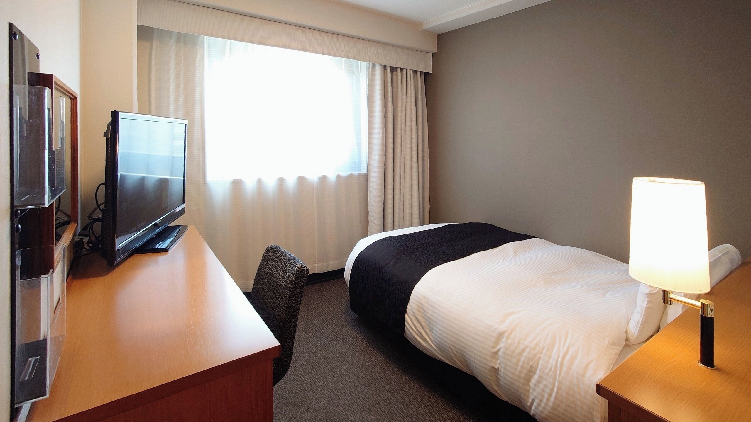 A hotel room with a bed, a desk with a television, a lamp, and a window with curtains.