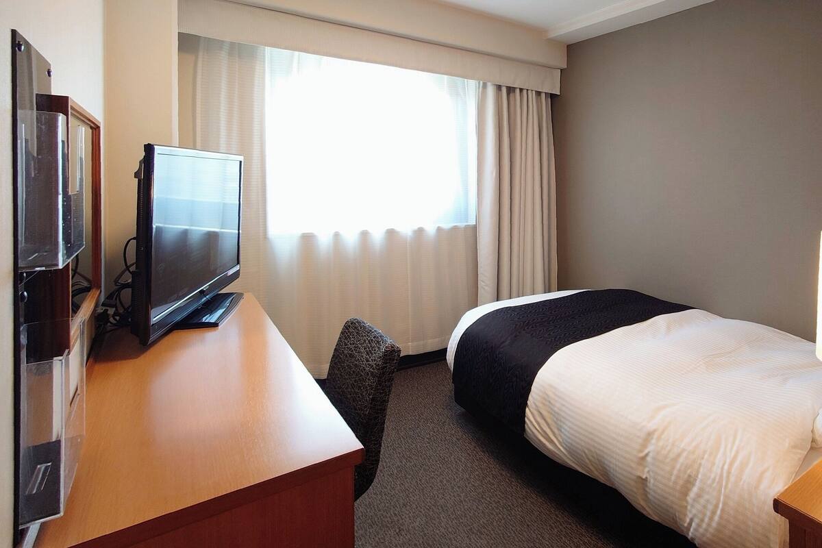 Ein Hotelzimmer mit einem Bett, einem Schreibtisch mit Fernseher, einer Lampe und einem Fenster mit Vorhängen.