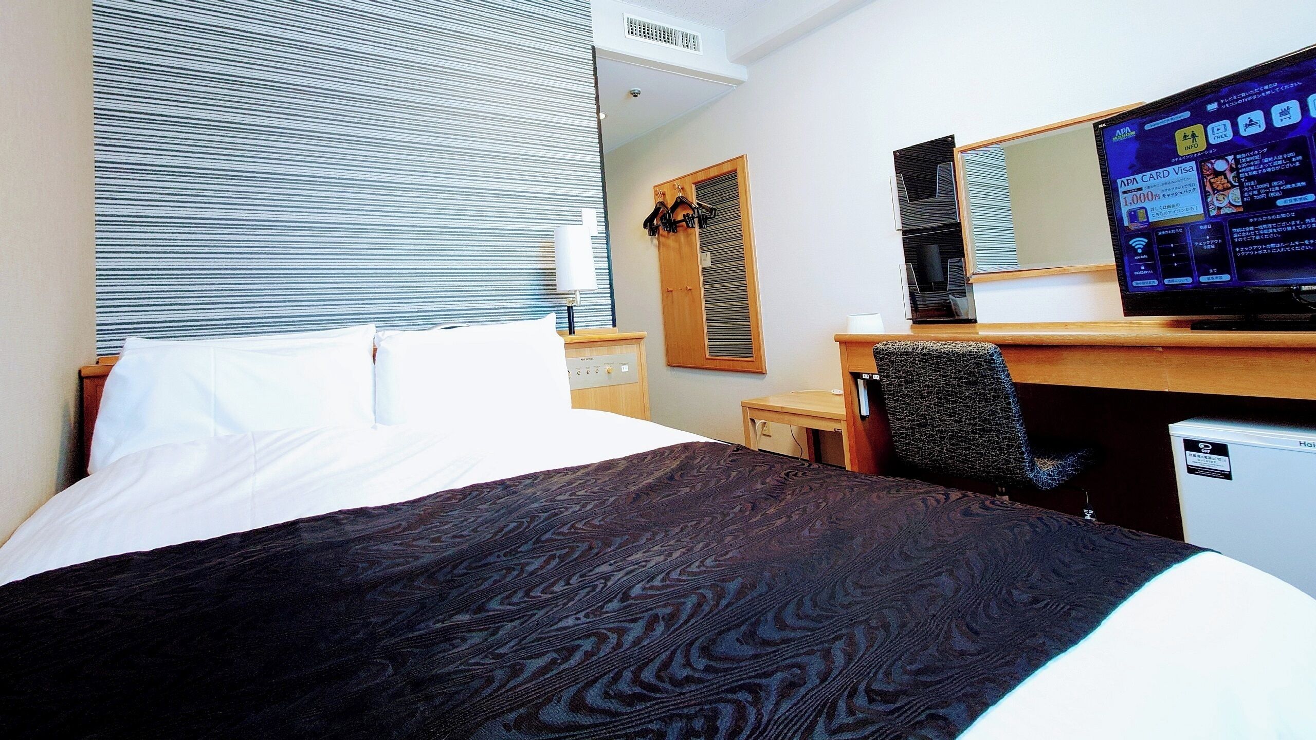 Quarto de hotel com cama, escrivaninha, televisão e janela com persianas.